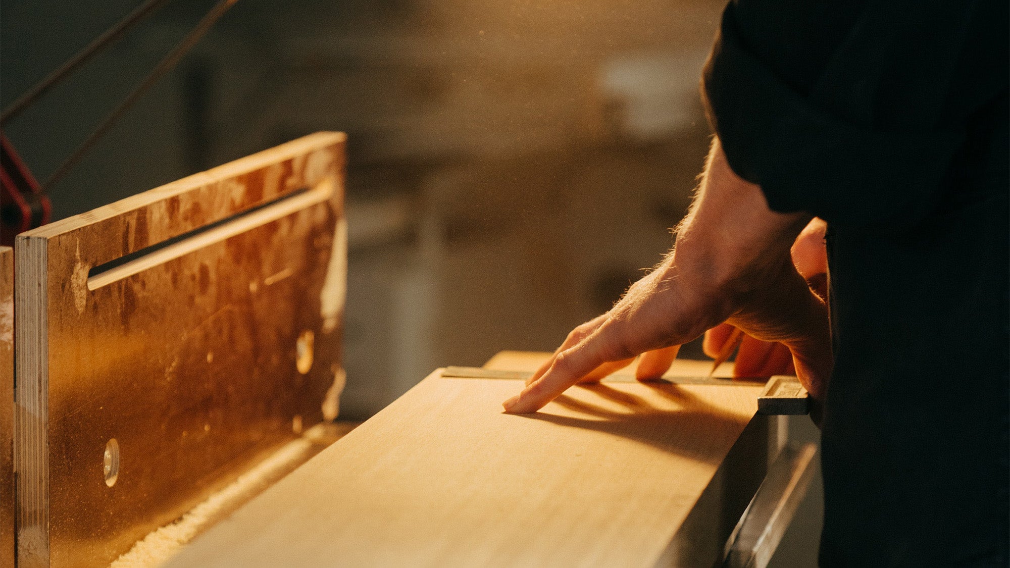 A closeup image of a man's hands measuring the width of a finished-edge slab of light colored wood on top of a work bench. He is using a small ruler and marking the wood with a pencil. The image has a warm tone and sawdust can be seen floating in the air.