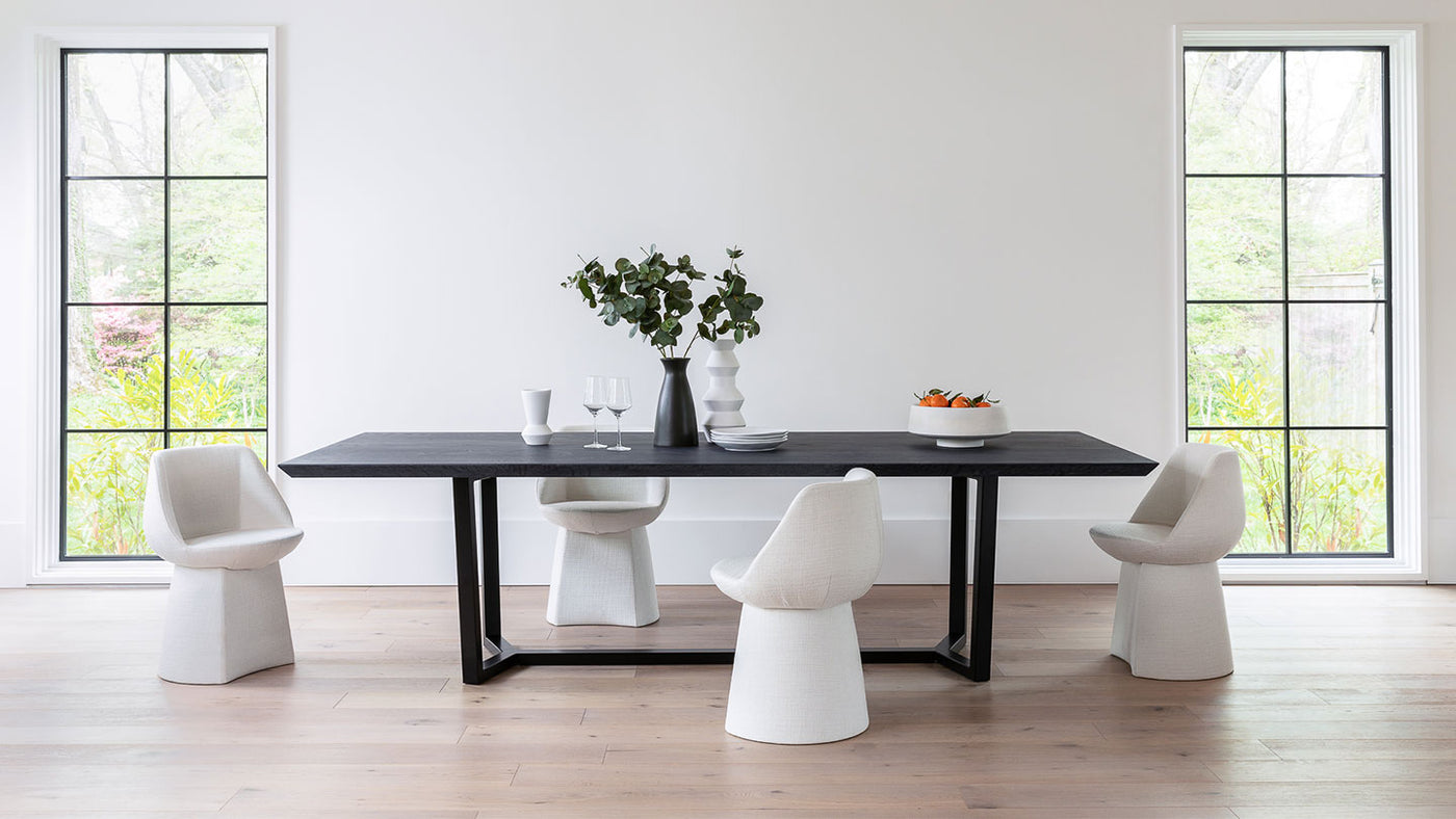 Black wood table with wood base surrounded by 4 white chairs in a white room on top of a light wood floor. The table is placed directly between two floor-to-ceiling windows with modern black framing. On top of the table sits a white decanter, two wine glasses, a black vase with eucalyptus branches, a white vase, a stack of 4 white plates, and a white fruit bowl with oranges
